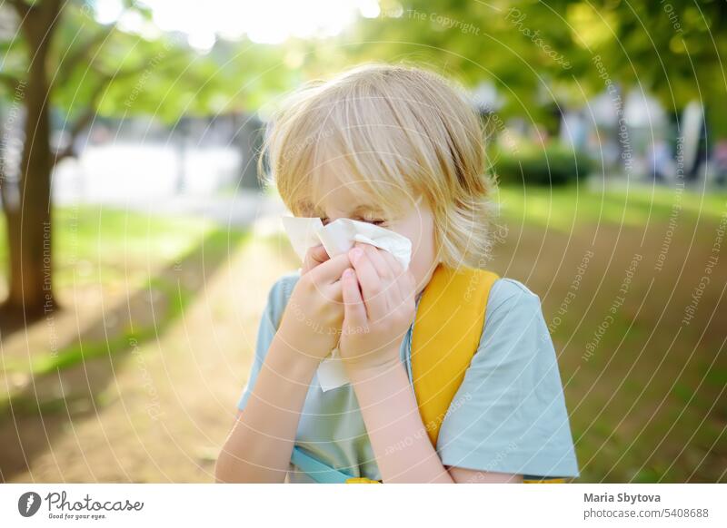 Ein Junge im Teenageralter niest und wischt sich die Nase mit einer Serviette ab, während er im Sommerpark spazieren geht. Grippesaison und Erkältungsschnupfen. Allergisches Kind.