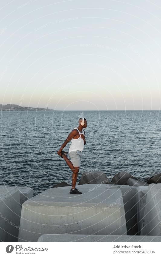 Schlanker schwarzer Mann, der sich beim Training die Beine an der Strandpromenade vertritt Läufer Hafengebiet Aufwärmen Kopfhörer Dehnung Energie Konzentration