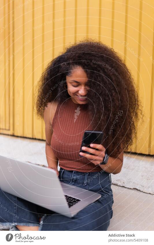 Fröhliche afroamerikanische selbständige Frau mit Laptop auf der Straße sitzend Smartphone freiberuflich krause Haare Großstadt Unternehmer ethnisch schwarz
