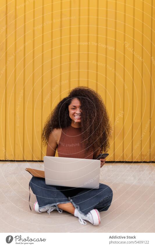 Fröhliche afroamerikanische selbständige Frau mit Laptop auf der Straße sitzend Smartphone freiberuflich krause Haare Großstadt Unternehmer ethnisch schwarz