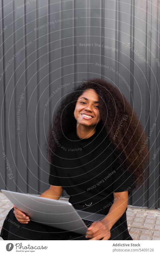 Fröhliche afroamerikanische selbständige Frau mit Laptop auf der Straße sitzend freiberuflich krause Haare Großstadt Unternehmer ethnisch schwarz Afroamerikaner