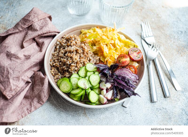 Teller mit leckerem Veggie-Gericht, Besteck und Gläser auf dem Tisch Vegetarier Buchweizen Gemüse Zwiebel Salatgurke Tomate Basilikum Schalen & Schüsseln