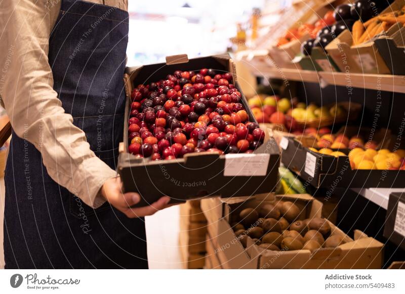 Anonymer männlicher Verkäufer zeigt Kiste mit Seilpflaumen im Supermarkt Mann Pflaume Frucht Lebensmittelgeschäft Gemüse manifestieren reif gesunde Ernährung