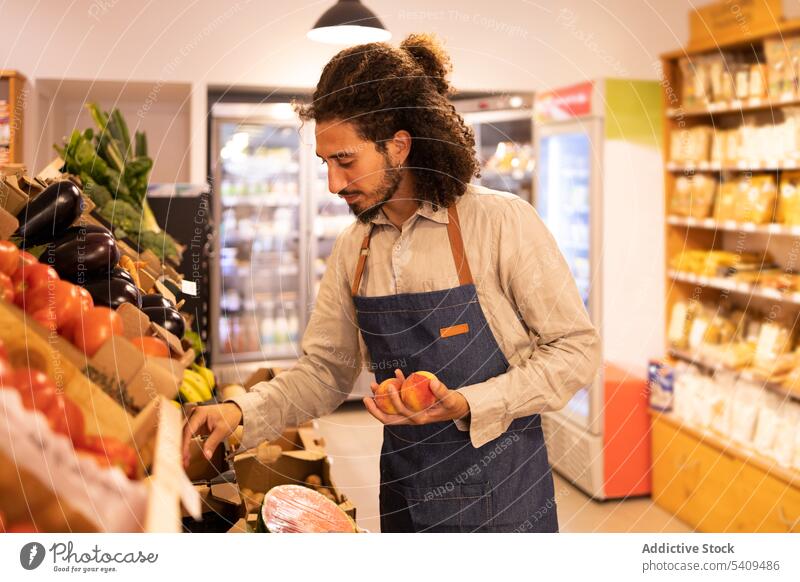 Ethnischer männlicher Verkäufer bei der Auswahl von Pfirsichen während der Arbeit im Supermarkt Mann Lebensmittelgeschäft Frucht wählen Abfertigungsschalter
