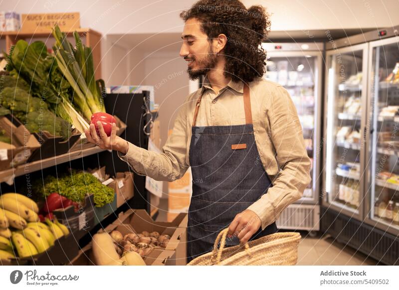 Lächelnder ethnischer Angestellter, der während seiner Arbeit im Lebensmittelladen Gemüse auswählt Mann Paprika Lebensmittelgeschäft wählen Supermarkt Laden