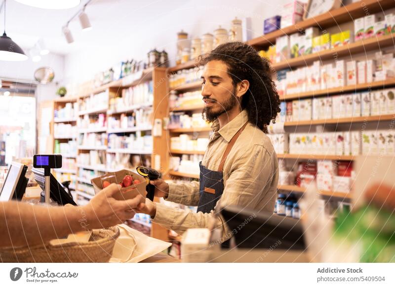 Junger ethnischer männlicher Kassierer scannt Produkte während der Arbeit in einem umweltfreundlichen Geschäft Mann Erdbeeren Verkäufer Frucht Supermarkt frisch