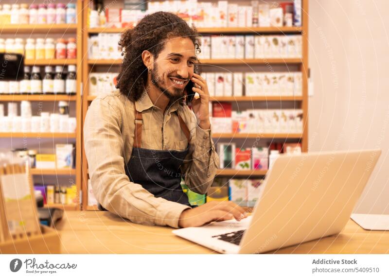 Lächelnder junger Mann, der in einem Geschäft mit seinem Smartphone spricht und auf einem Laptop surft reden positiv Kommunizieren Apparatur Drogerie Browsen