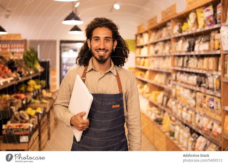 Fröhlicher ethnischer Mann mit Laptop in einem Lebensmittelladen Lächeln Werkstatt Laden positiv Arbeit Schürze Job Apparatur Glück lässig Vollbart