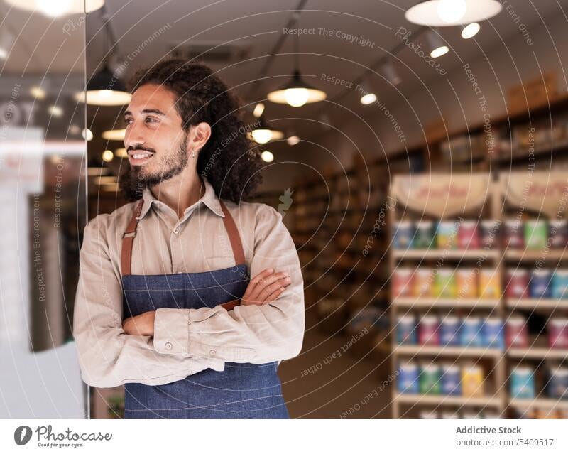 Fröhlicher Mann, der vor einem beleuchteten Supermarkt mit Waren steht Lächeln selbstbewusst Porträt Vollbart positiv heiter Glück Schürze Arbeit freundlich