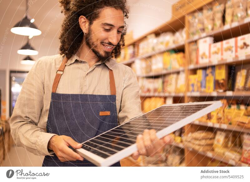 Fröhlicher ethnischer Verkäufer mit tragbarem Solarpanel im Laden Mann Supermarkt Werkstatt Glück heiter solar Panel Arbeiter positiv Kauf modern Gerät Käufer