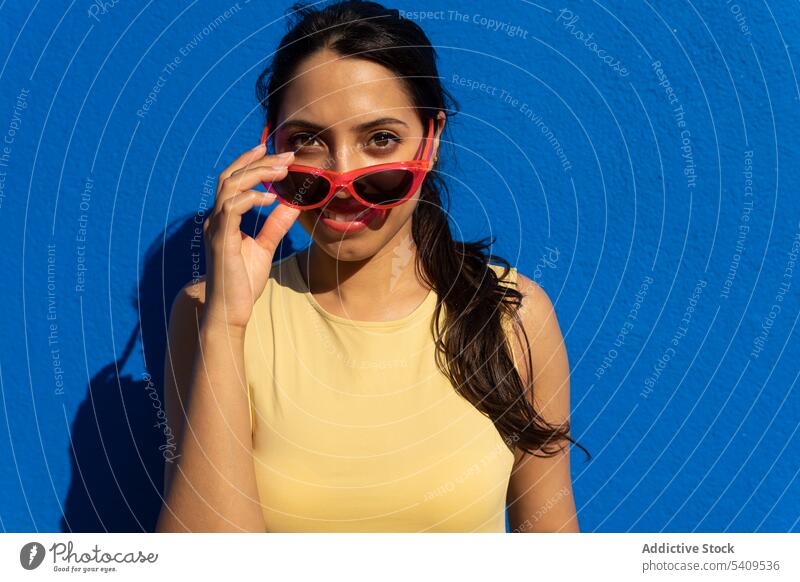 Glückliche junge ethnische Frau mit Sonnenbrille steht im Licht gegen blaue Wand Lächeln Schatten Porträt Raum Vorschein positiv charmant heiter Inder attraktiv