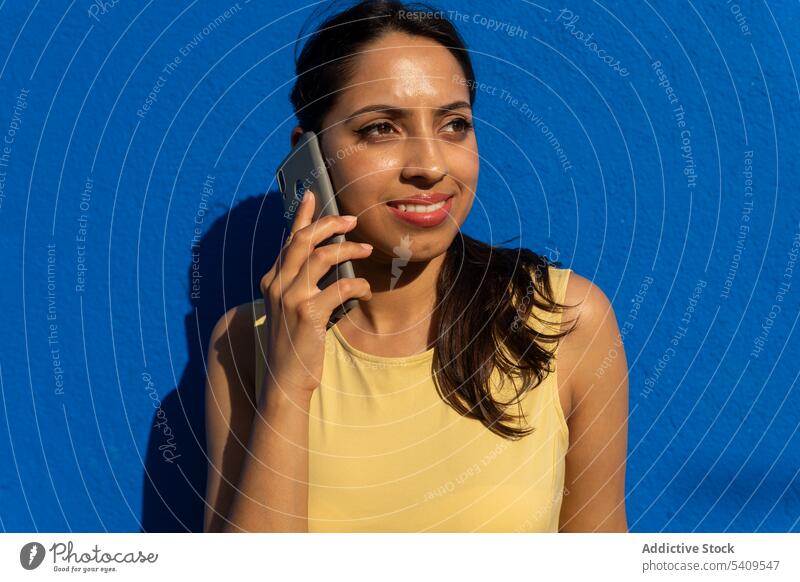 Lächelnde junge ethnische Frau, die mit einem Smartphone vor blauem Hintergrund spricht sprechen Porträt Schatten lange Haare Kommunizieren Glück positiv Mobile