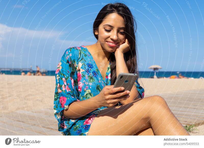 Lächelnde junge ethnische Frau sitzt am Strand und surft auf ihrem Smartphone vor blauem Himmel Porträt benutzend Glück positiv lange Haare Blauer Himmel
