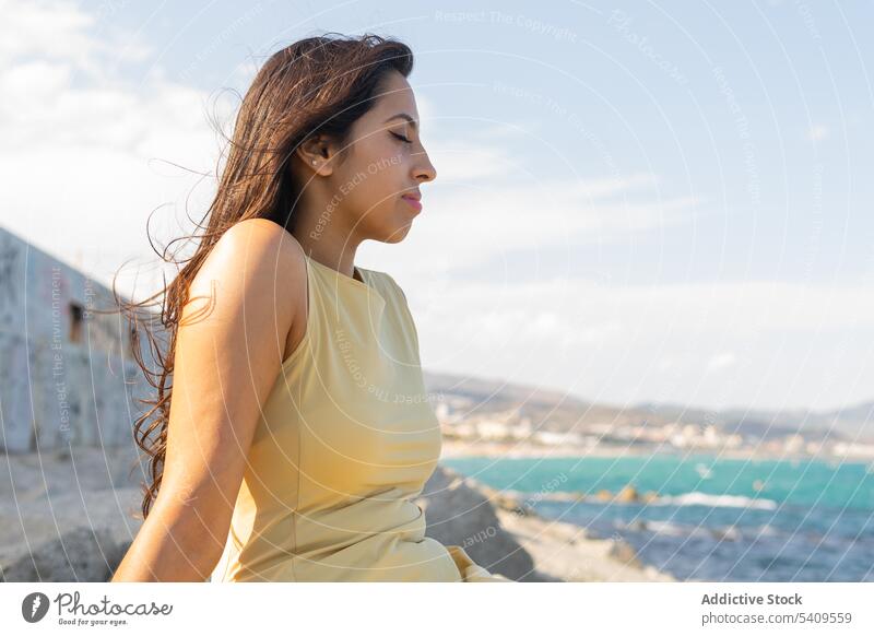 Junge ethnische Frau sitzt am Strand im Tageslicht mit geschlossenen Augen in der Nähe von verschwommenem Meerwasser Lächeln Augen geschlossen Küste Natur