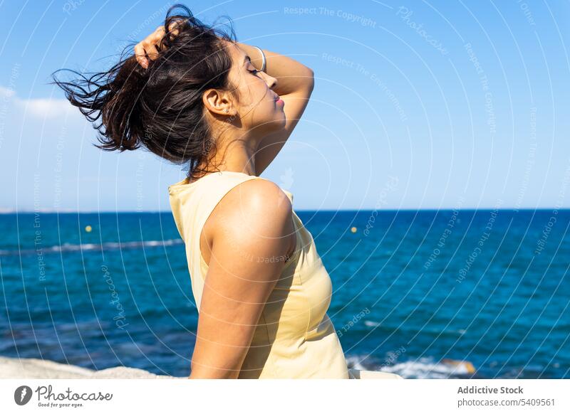 Junge ethnische Frau sitzt am Strand im Tageslicht mit geschlossenen Augen in der Nähe von verschwommenem Meerwasser Augen geschlossen Küste Natur lange Haare