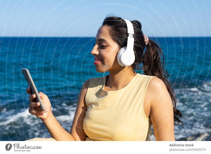 Indische Frau mit Kopfhörern und Smartphone am Meer benutzend zuhören Browsen Musik Lächeln MEER Strand genießen Meeresufer ruhen heiter Apparatur Gerät Mobile