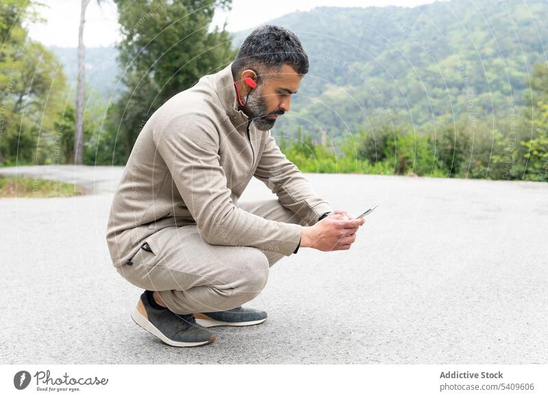 Fokussierter ethnischer Mann mit TWS-Kopfhörern, der auf der Straße sitzt und sein Smartphone benutzt Sportler tws benutzend Ohrstöpsel Nachricht online