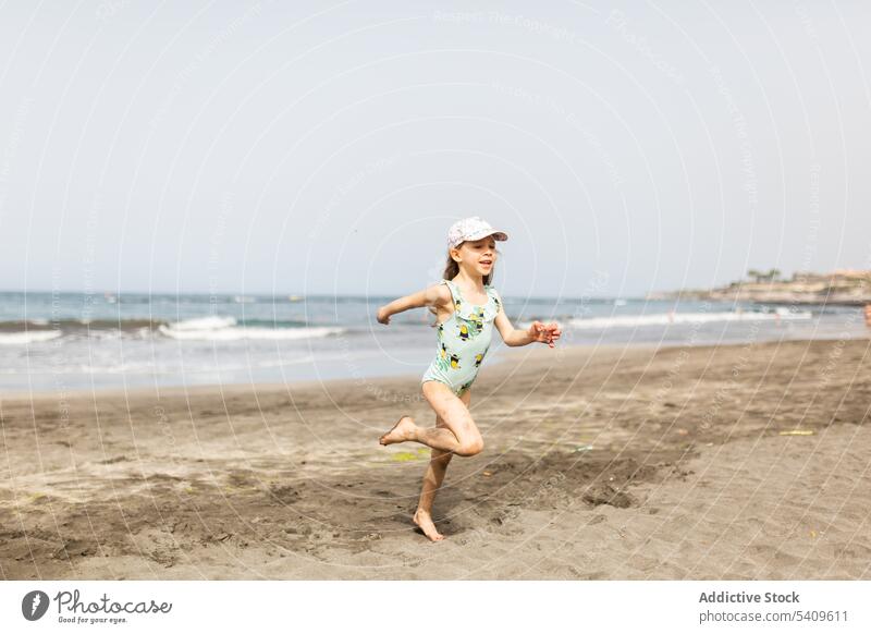 Glückliches Kind in Badekleidung und Mütze läuft am Sandstrand laufen Strand nass Barfuß Lächeln Meeresufer sorgenfrei Sommer Mädchen Urlaub winken