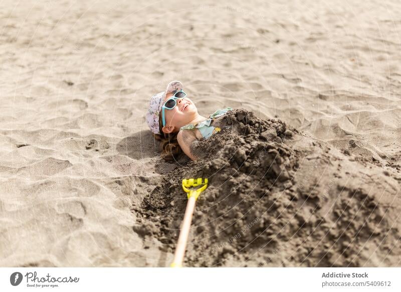 Glückliches Kind mit Mütze und Sonnenbrille auf dem Boden liegend mit nassem Sand auf dem Körper Strand sorgenfrei Lügen Spaß haben spielerisch Lächeln Sommer