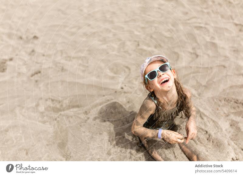Fröhliches Kind mit Sonnenbrille am Sandstrand sitzend mit Mütze im Tageslicht genießen Urlaub Strand Lächeln Verschlussdeckel Badebekleidung Sommer Mädchen