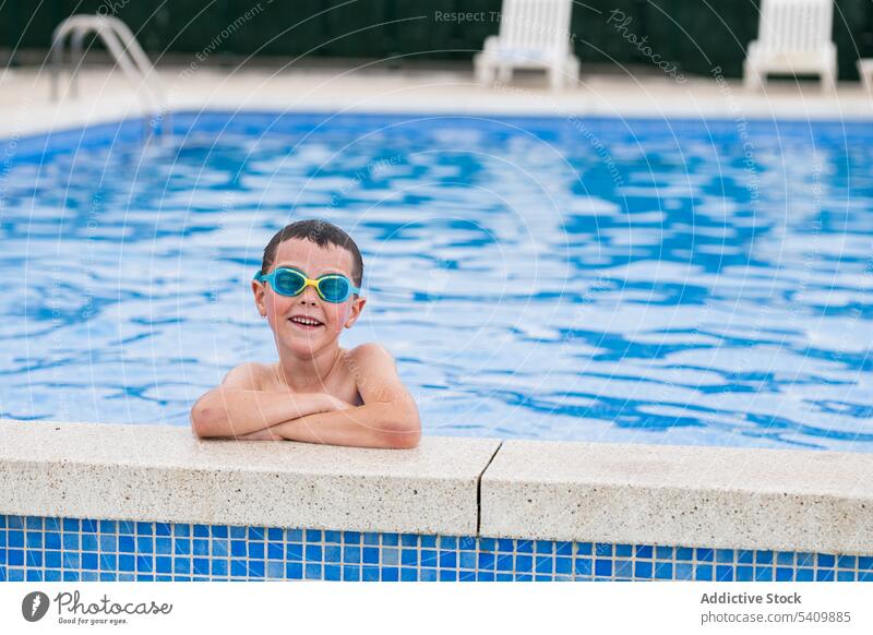 Glückliches Kind mit Schwimmbrille im Schwimmbad stehend bei Tageslicht Lächeln Schutzbrille Pool nasses Haar Wasser genießen froh die Hände gekreuzt Beckenrand