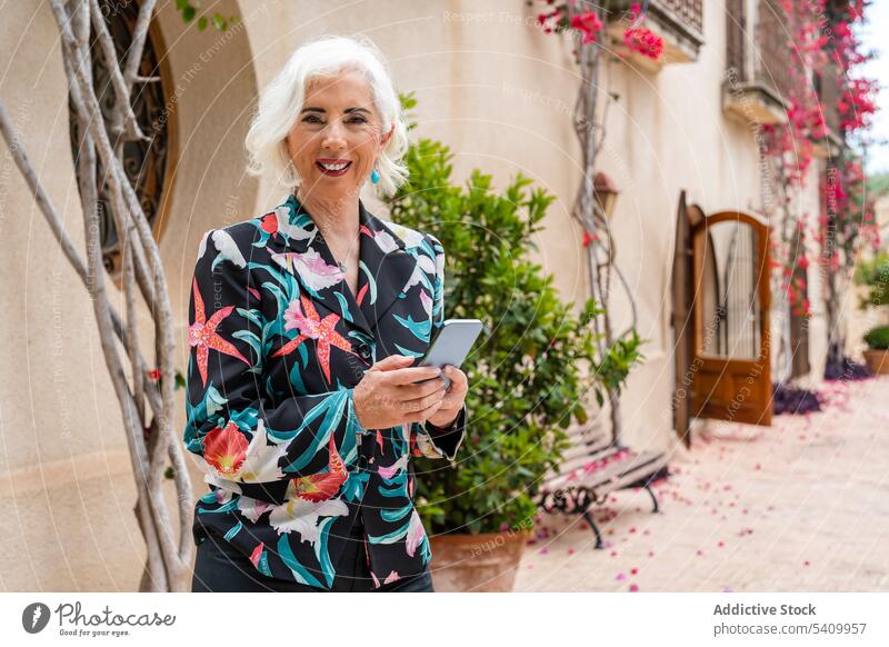 Lächelnde ältere Frau, die auf dem Gehweg steht und ein Smartphone benutzt benutzend Gebäude Bürgersteig Türöffnung online Pflanze Außenseite Senior Browsen