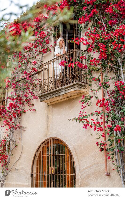 Ältere Frau steht auf Balkon mit Eisenzaun in der Nähe von grünen Blättern Baum mit Blumen Blütezeit Gebäude allein Zaun Haus Außenseite Windstille Senior
