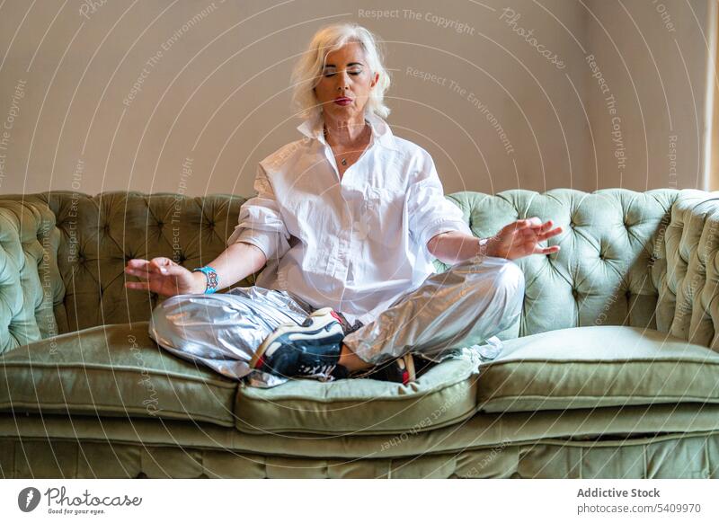 Ältere Frau sitzt auf dem Sofa mit gekreuzten Beinen und macht Padmasana Pose Yoga meditieren Lotos Mudra Beine gekreuzt üben Augen geschlossen Achtsamkeit