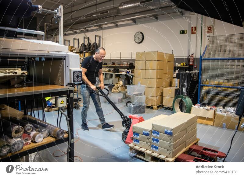Starker Mann mittleren Alters, der in einer Fabrik Material mit einem Palettenwagenheber zieht Arbeiter Lagerhalle Inszenierung professionell ziehen Wagenheber