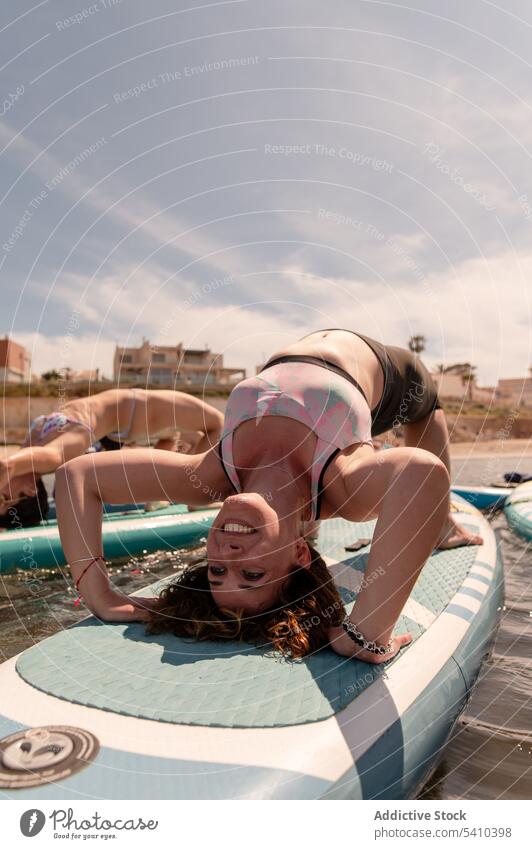 Unbekannte Frauen machen Yoga auf dem Paddleboard bei Tageslicht Freundin üben Halbrad Paddelbrett beweglich Gleichgewicht Badebekleidung Meerwasser jung wolkig
