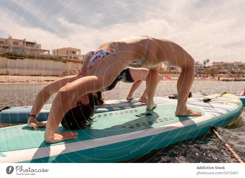 Unbekannte Frauen machen Yoga auf dem Paddleboard bei Tageslicht Freundin üben Halbrad Paddelbrett beweglich Gleichgewicht Badebekleidung Meerwasser jung wolkig