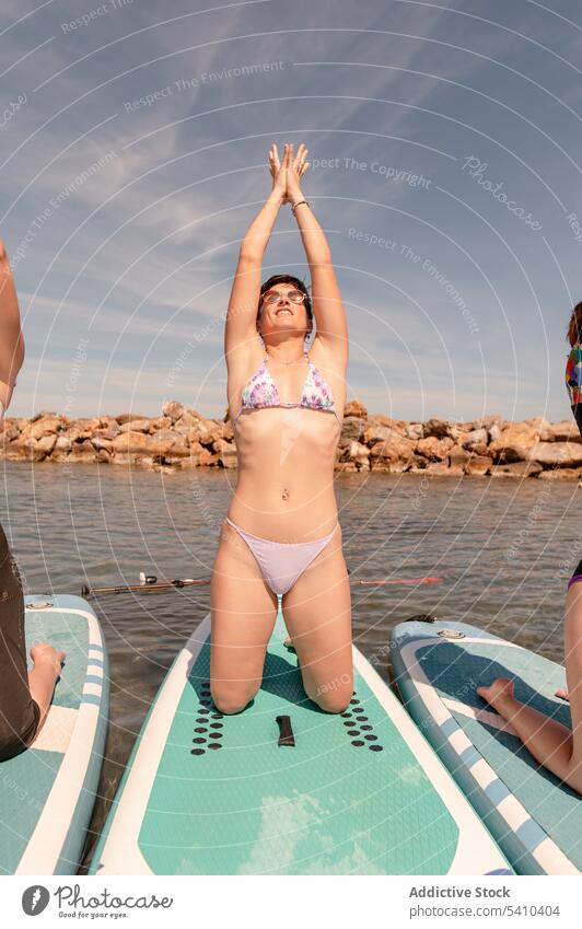 Junge Frau in Yoga-Pose auf dem Paddelbrett Freund Augen geschlossen Mudra Dehnung knien Berg mit erhobenen Armen jung Sommer wolkig Sonnenbrille Blauer Himmel