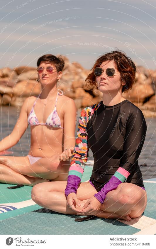Junge Frauen sitzen in Padmasana-Pose auf Paddelbrettern Yoga Lotos meditieren üben Windstille Sonnenbrille Konzentration jung Sommer Erholung Zen Urlaub Asana
