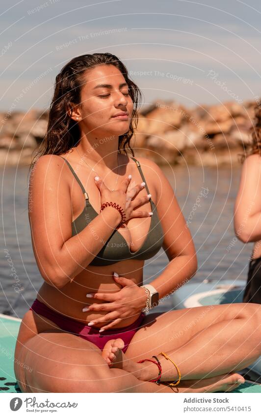 Ruhige Frau in Badehose atmet beim SUP-Yoga Sup-Yoga Paddelbrett Zusatzplatine meditieren Gleichgewicht Zen Atem Seeküste üben Windstille Harmonie Pose Wellness