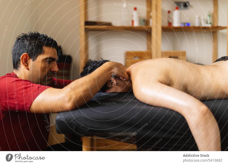 Ethnischer erwachsener Masseur massiert die Schultern einer liegenden anonymen Kundin im Salon Frau Klient Massage oben ohne Verfahren Wellness sinnlich