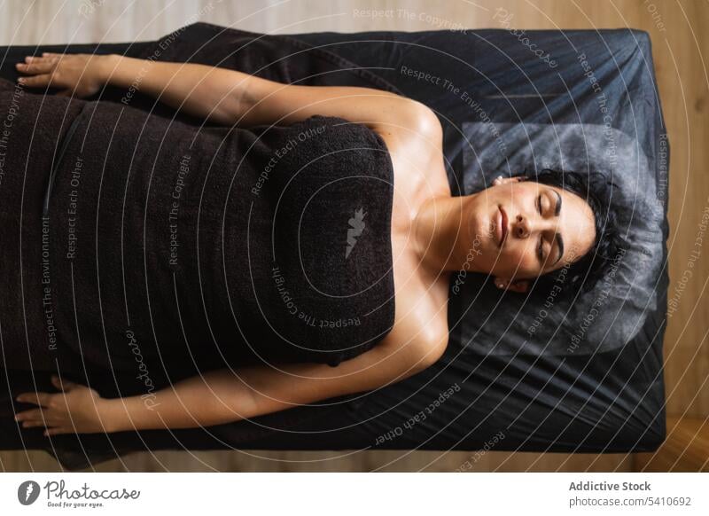Junge ethnische Frau liegt auf einem bequemen Massagebett in einem Salon am Tag Bett sich[Akk] entspannen Verfahren Hautpflege Handtuch Augen geschlossen