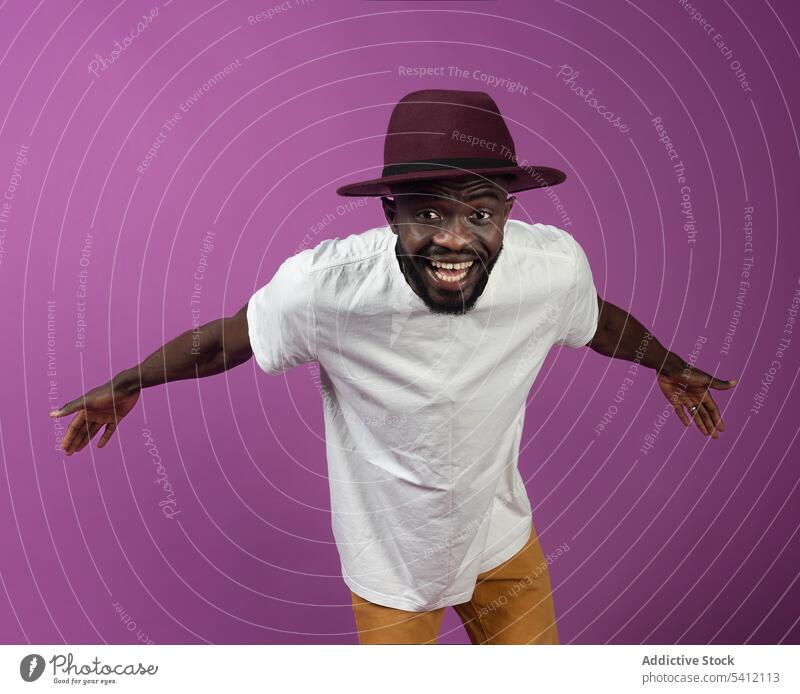 Fröhlicher schwarzer Mann mit Hut steht mit ausgestreckten Armen in der Nähe einer lila Wand Lächeln Glück aufgeregt freundlich ausdehnen Freiheit positiv froh
