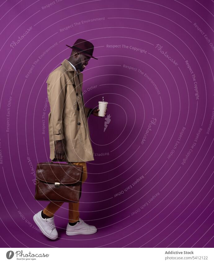 Glücklicher junger schwarzer Mann mit Aktentasche und Kaffeebecher zum Mitnehmen geht in der Nähe einer lila Wand Lächeln Spaziergang Heißgetränk Imbissbude