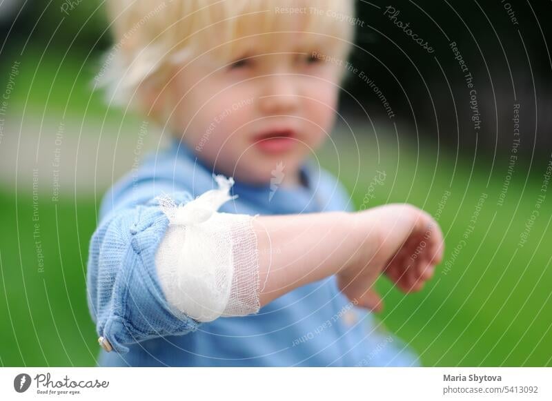Niedlicher kleiner Junge schaut auf seinen Ellbogen mit angelegtem Verband. Erste Hilfe am Kind. bandagieren Wunde blaue Flecken wehtun medizinisch Gaze