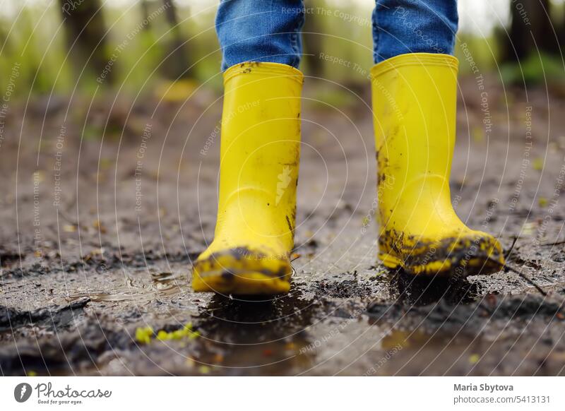 Verschmitztes Vorschulkind in gelben Regenstiefeln, das in eine große nasse Schlammpfütze springt. Kind spielt und hat Spaß. Spiele im Freien springend Stiefel