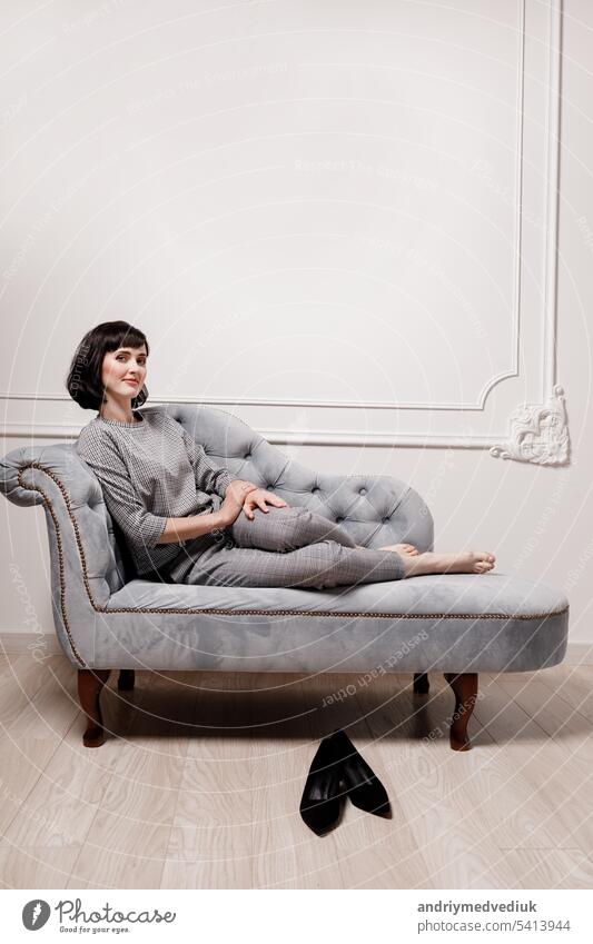 Porträt einer positiven jungen brünetten Frau, die barfuß auf einem grauen Sofa mit ausgestreckten Beinen in einem grau karierten Anzug sitzt und in die Kamera schaut. Eine Geschäftsfrau zieht ihre Schuhe aus und ruht sich nach der Arbeit aus.