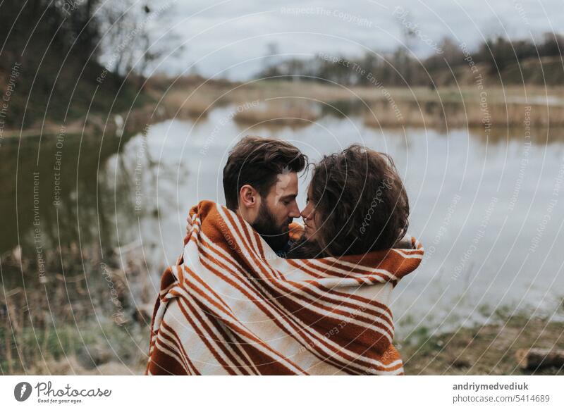 Zwei kaukasische Liebende in Decke eingewickelt am See. Junges Paar umarmt sich an einem Herbsttag im Freien. Ein bärtiger Mann und eine lockige Frau in der Liebe. Valentinstag. Konzept der Liebe und Familie.