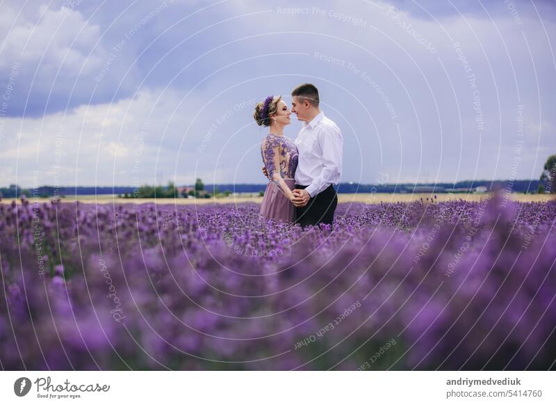 Junges verliebtes Paar, das sich umarmt und in einem Lavendelfeld am Sommertag spazieren geht. Mädchen in einem luxuriösen lila Kleid und mit Frisur Liebe