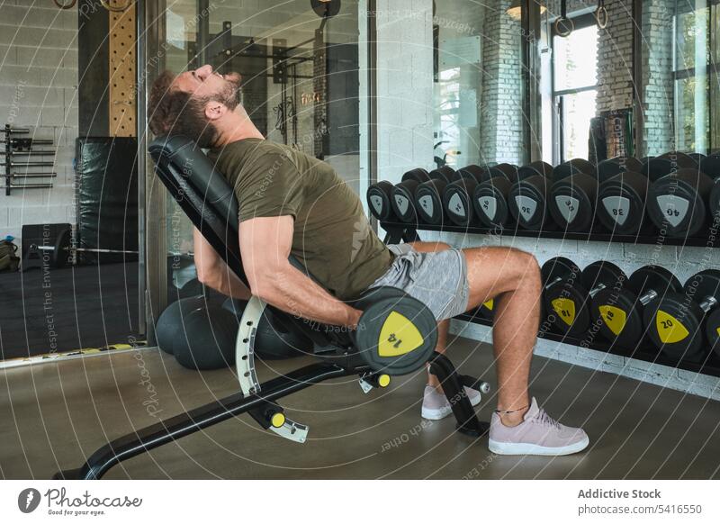 Starker Mann beim Training mit Hanteln im Fitnessstudio Heben Bank Sitzen Übung Sport männlich muskulös stark Athlet anstrengen physisch Gesundheit Wellness