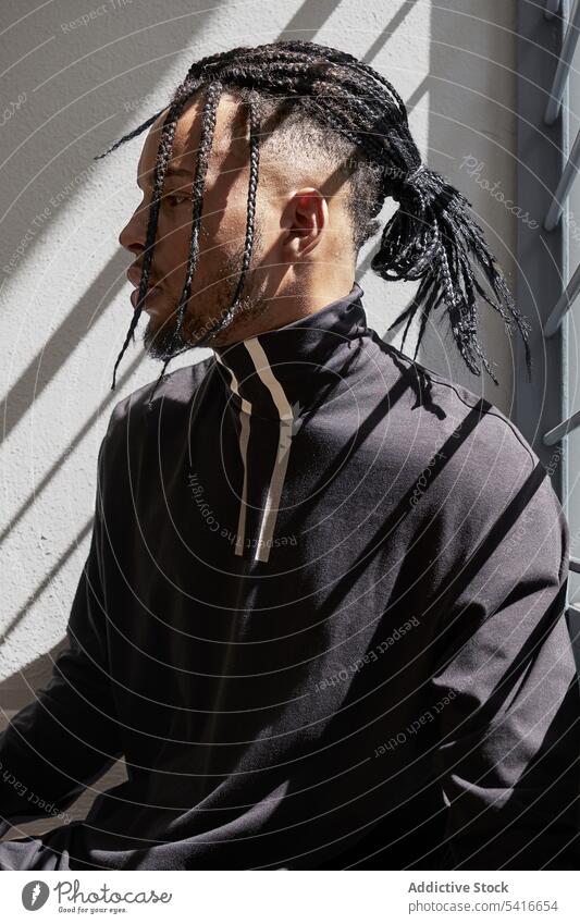 Stilvoller ethnischer Mann auf Geländer sitzend Geflecht Frisur Zöpfe cornrows trendy Treppenhaus Reling jung männlich Afroamerikaner Person gutaussehend