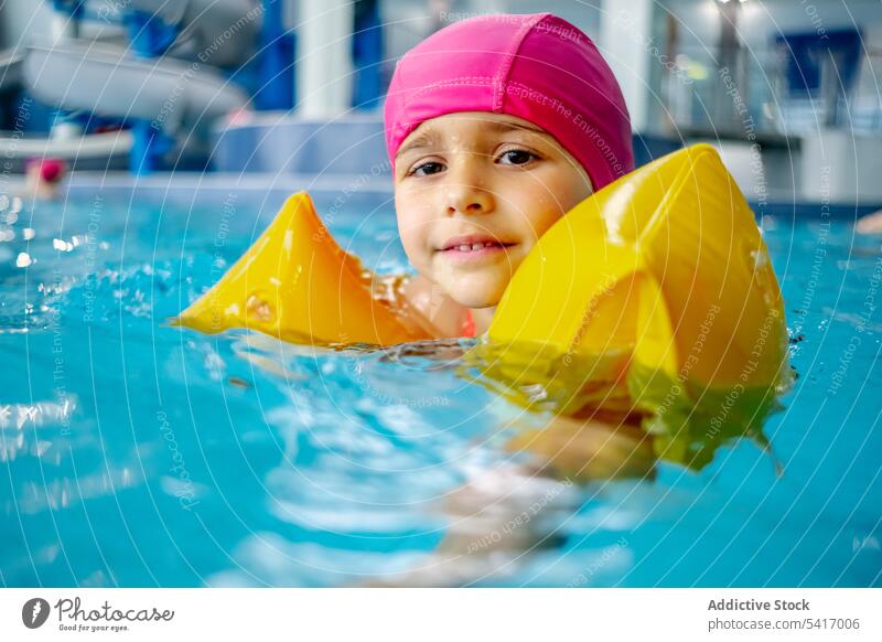 Fröhliches ethnisches Mädchen mit Hut, lächelnd und schwimmend im Pool im Wasserpark Schwimmsport Lächeln Park Freizeit Kind Spaß zwei Finger Frieden Sieg