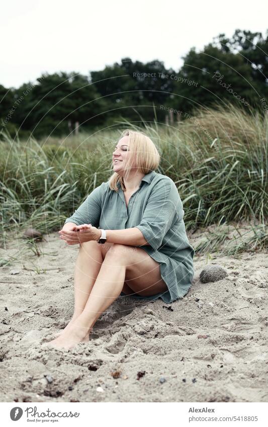 Portrait einer schönen, blonden Frau die im Sand am Strand sitzt und sich freut langhaarig blonde Haare Freude Lächeln Dünengras Schönheit entspannt relaxed