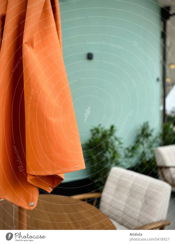 Geschlossener orangener Sonnenschirm mit Sitzmöbeln vor mintgrüner Wand geschlossen Polsterung Terrasse Außenaufnahme Fassade Farbkontrast Grünpflanze