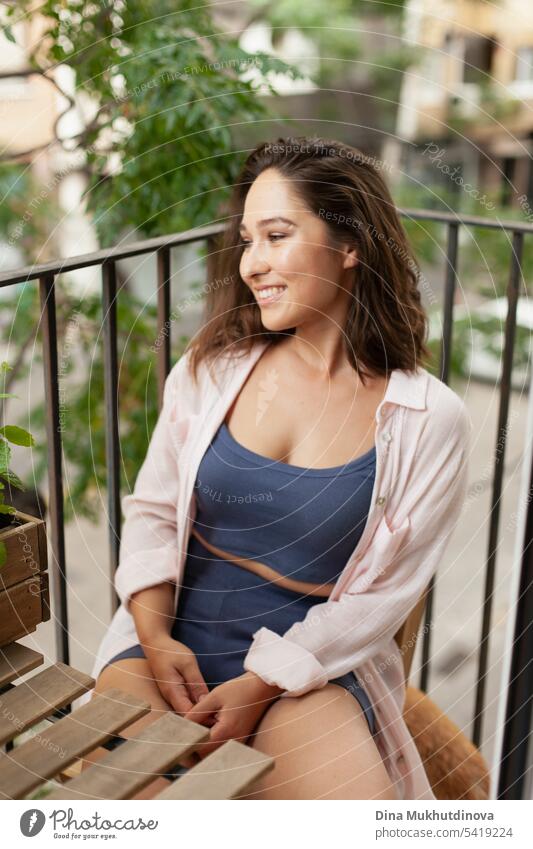 Schöne Frau sitzt auf einem Holzstuhl auf dem Balkon. Glückliche Brünette lächelnd. Pflanzen und Blumen Gartenarbeit Hobby. Balkonpflanze Balkondekoration