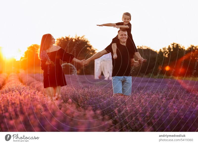 glücklicher Familientag. Junge Mutter und Vater trägt auf den Schultern Tochter in Lavendelfeld auf Sonnenuntergang. Papa, Mama und Kind Mädchen haben Spaß auf die Natur im Sommer. Konzept der freundlichen Familie. Familie Blick